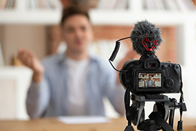Image of camera with external microphone pointed at a young person who is speaking several feet away. The speaker is blurred while the camera and the recording are crispy and visible.