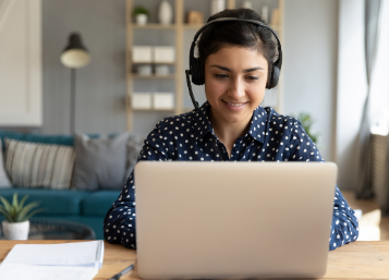 Image of person with headphones on in a living room sitting at a desk looking at a laptop screen with a notebook on the desk.