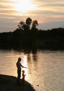 A photograph of a father and son beside a lake who are silhouetted as the sun sets behind them