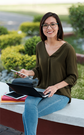 A photograph of a student sitting outside in a landscaped setting with notebooks beside her, a cellphone in one hand and a laptop computer on her lap.