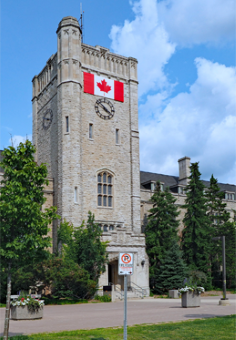 A photograph of the Johnston Hall clock tower at the University if Guelph on a sunny day. There is a Canadian flag hanging on the tower above the clock.