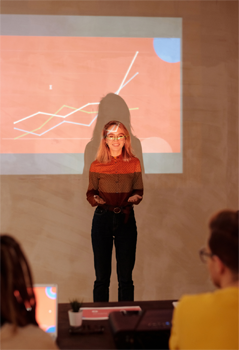 A photograph of a woman at a podium presenting to a group of seated people.