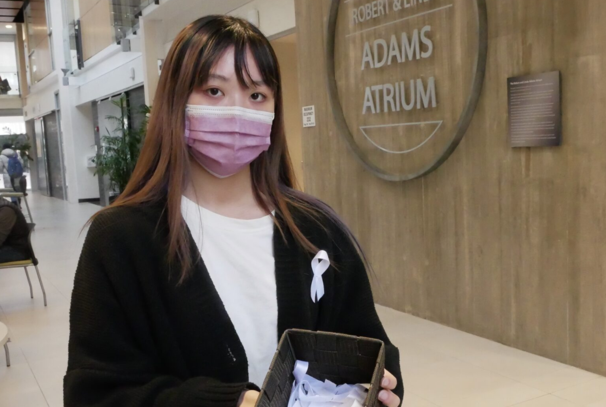 Grace Ly wearing mask while standing in the Adams Atrium and holding basket of white ribbons