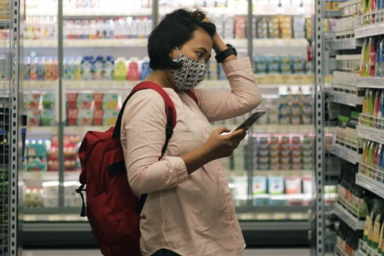 A person wearing a backpack and holding their smartphone is standing in a grocery store aisle looking as though she is struggling to make a decision about what food items to purchase