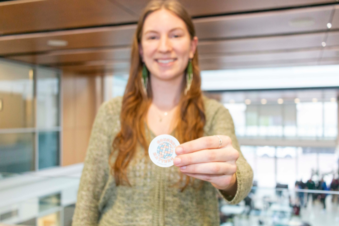 Student smiling and holding a sticker to camera which is in focus.